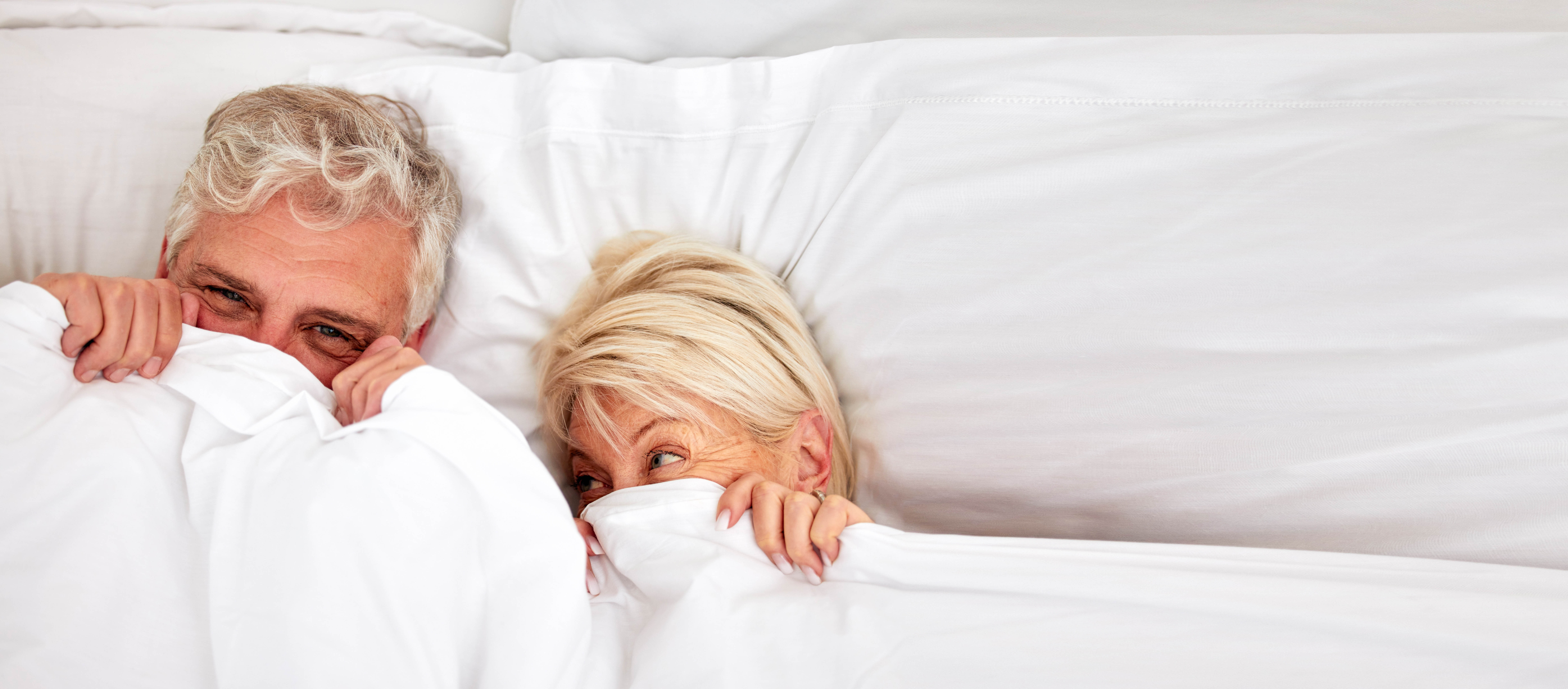 Older couple laying in bed with the covers pulled up over their noses, and being held in place
